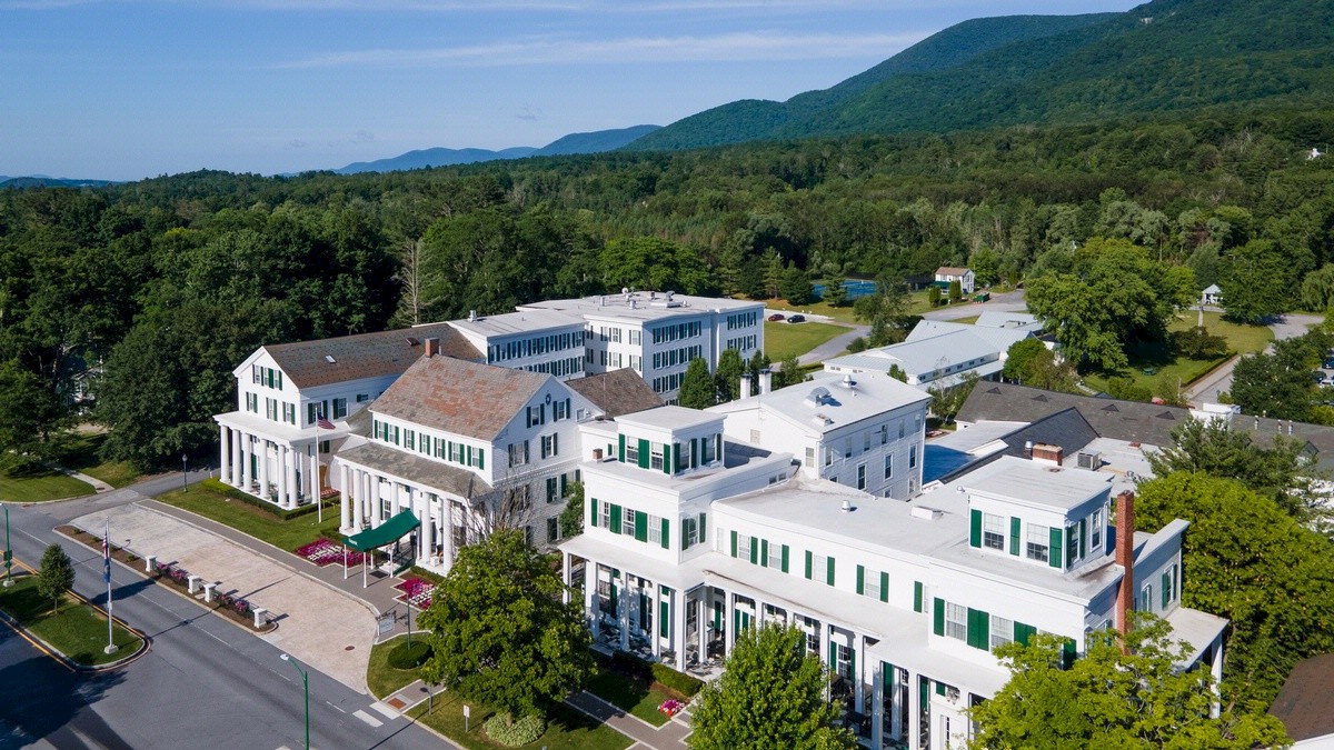 An aerial view of a large building complex with white facades, green shutters, and surrounded by greenery and mountains in the background.