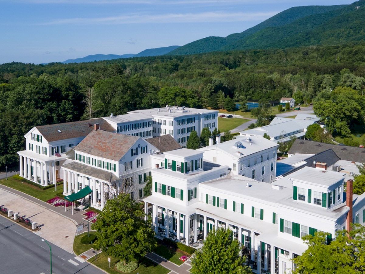 An aerial view of a large building complex with white facades, green shutters, and surrounded by greenery and mountains in the background.