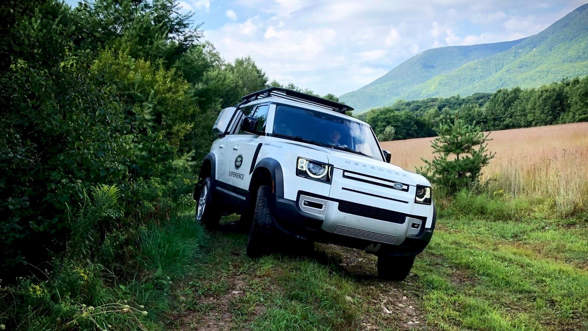 A white off-road vehicle parked on a grassy path next to thick greenery, with mountains and a cloudy blue sky in the background.