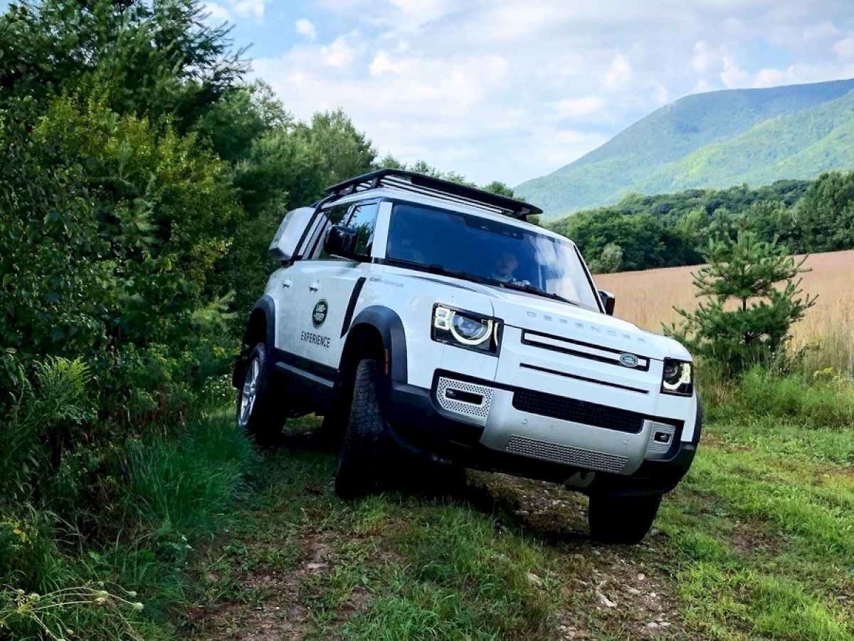 A white off-road vehicle parked on a grassy path next to thick greenery, with mountains and a cloudy blue sky in the background.