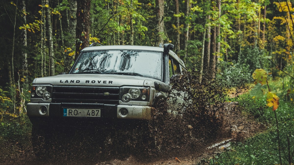 A white Land Rover is driving through a muddy, forested trail, splashing mud around as it navigates the path surrounded by greenery and tall trees.