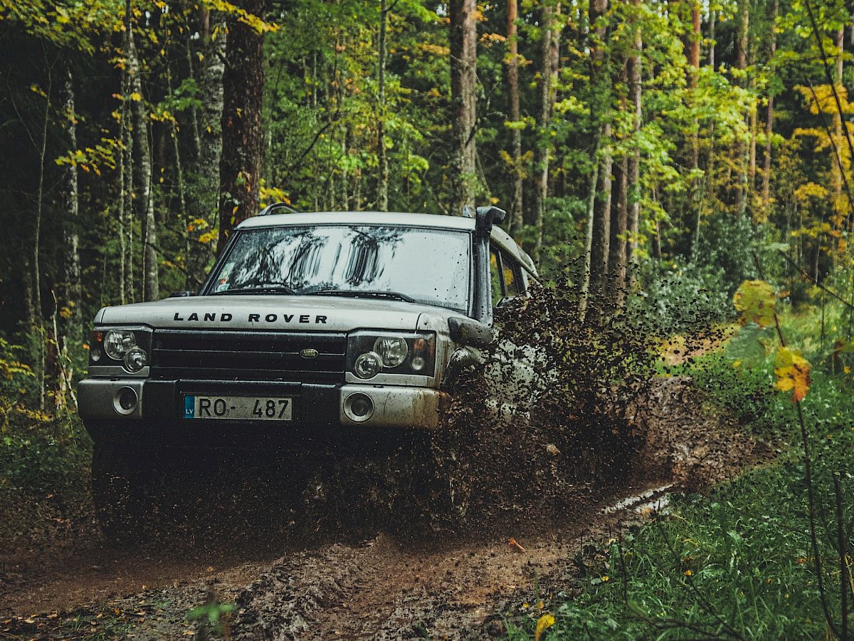 A white Land Rover is driving through a muddy, forested trail, splashing mud around as it navigates the path surrounded by greenery and tall trees.