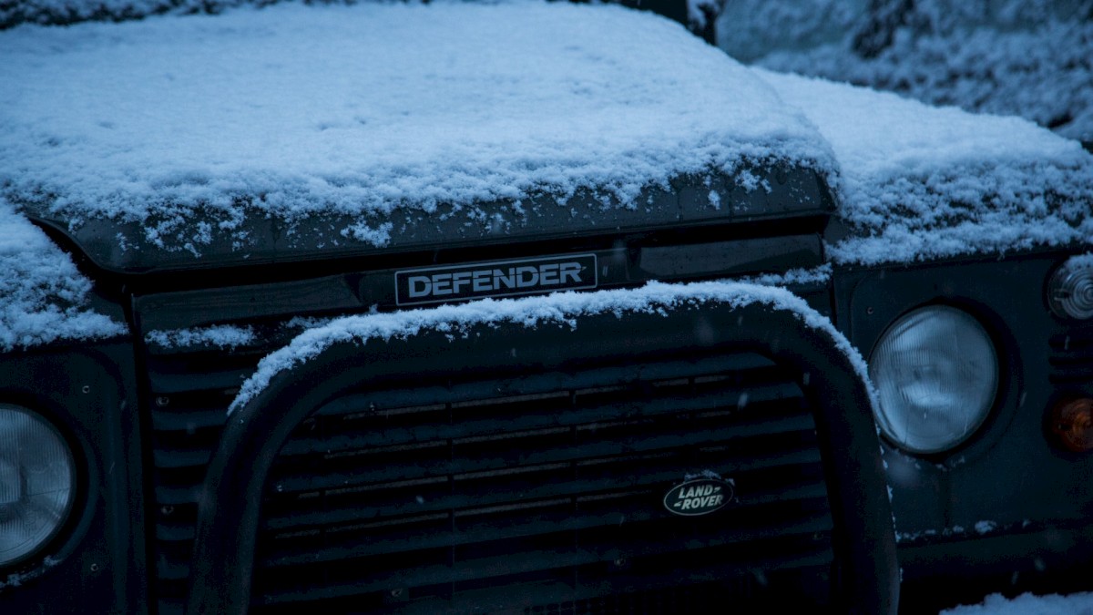 A snow-covered Land Rover Defender is pictured, highlighting the iconic front grille and headlight in a winter setting, partially obscured by snow.