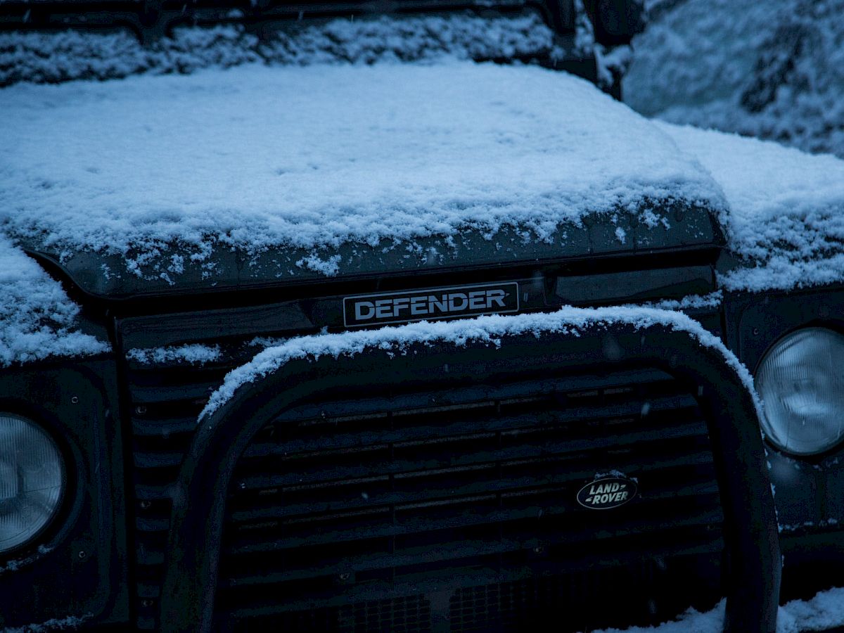 A snow-covered Land Rover Defender is pictured, highlighting the iconic front grille and headlight in a winter setting, partially obscured by snow.