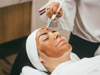 A person wearing a headband receives a facial treatment, with a brush applying a brown mask on their face while they relax.