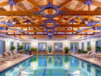 The image shows an indoor pool area with wooden ceiling beams, lounge chairs, plants, and tables with chairs. The room has a relaxed and inviting ambiance.
