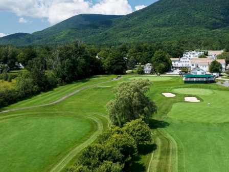 A scenic golf course with lush green fairways, a few sand bunkers, trees, residential buildings, and mountains in the background on a clear day.