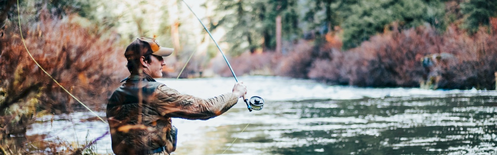 A person is fly fishing in a river surrounded by trees, wearing outdoor gear, and casting a line into the water.