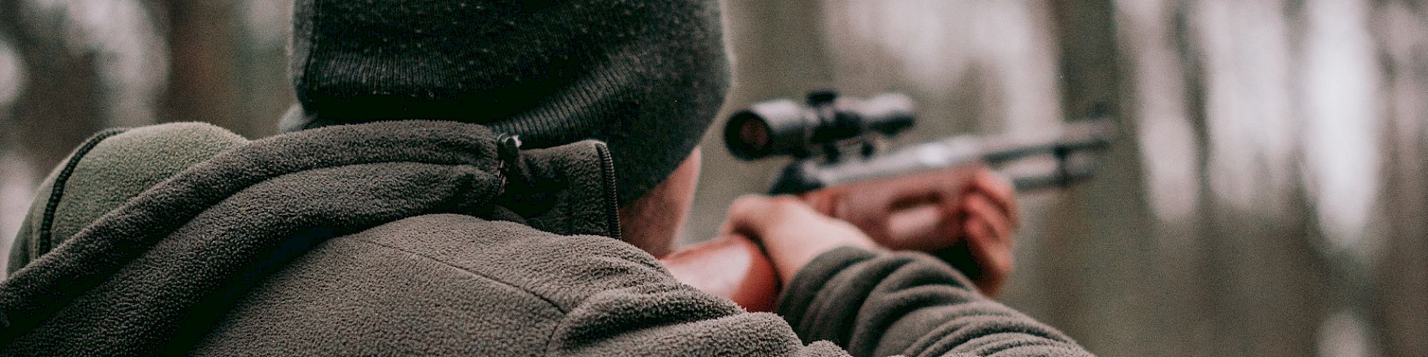 A person wearing a beanie and jacket is aiming a rifle in a wooded area, surrounded by trees on an overcast day.