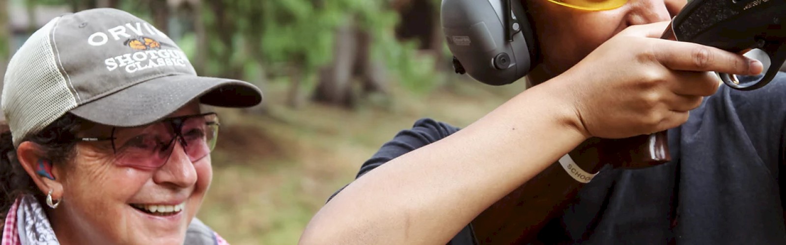 Two people are outdoors, one is aiming a gun wearing protective gear, guided by someone smiling next to them.