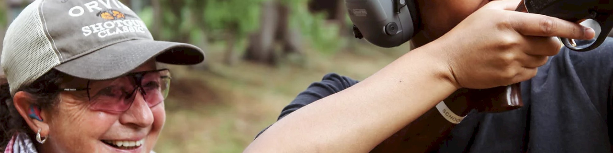 Two people are outdoors, one is aiming a gun wearing protective gear, guided by someone smiling next to them.