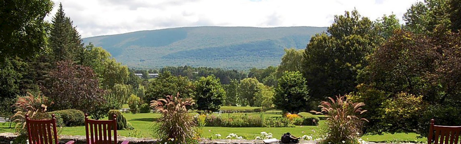 A scenic landscape features a manicured garden with chairs in the foreground, trees, and a mountainous backdrop under a partly cloudy sky.