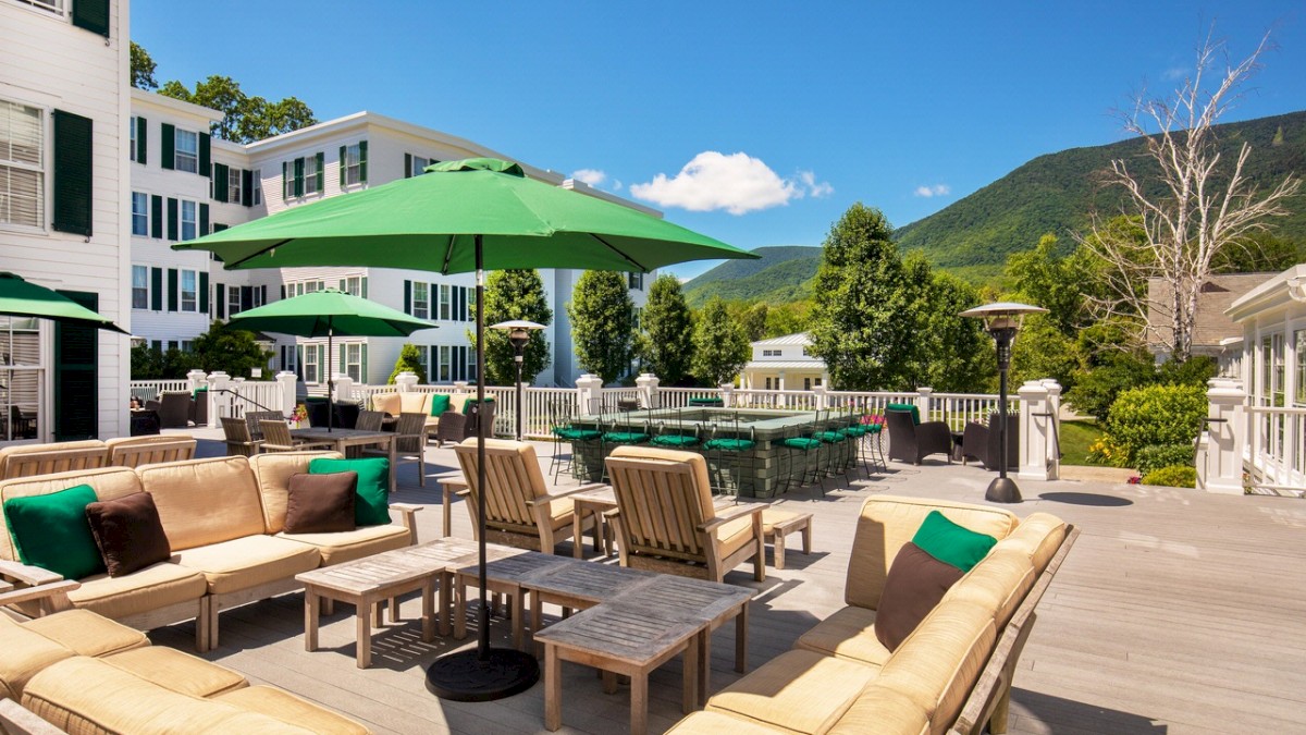 An outdoor seating area with cushioned furniture, green umbrellas, and scenic mountain views in the background under a clear blue sky.