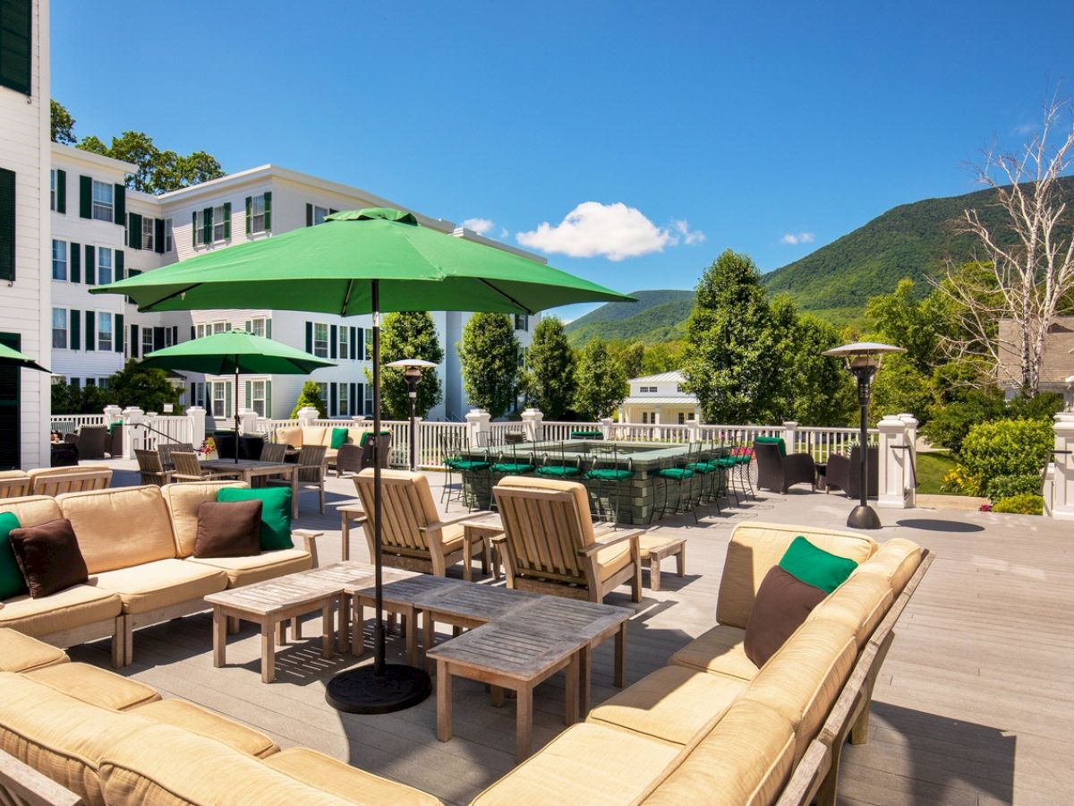 An outdoor seating area with cushioned furniture, green umbrellas, and scenic mountain views in the background under a clear blue sky.
