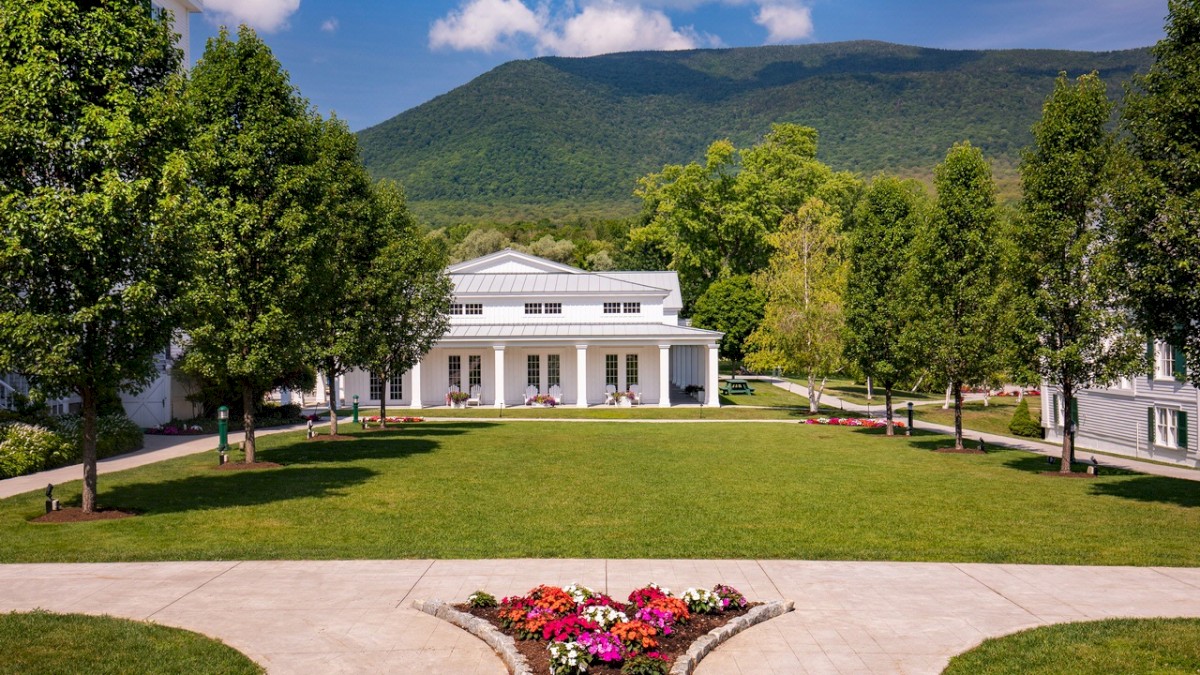 The image shows a manicured lawn with flower beds, surrounded by trees and white buildings, set against a mountainous backdrop under a blue sky.