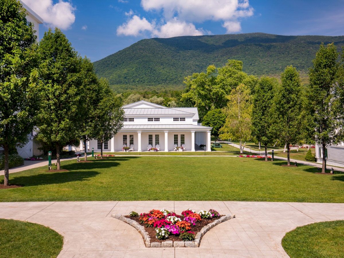 The image shows a manicured lawn with flower beds, surrounded by trees and white buildings, set against a mountainous backdrop under a blue sky.