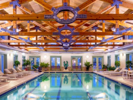 The image shows an indoor swimming pool with lounge chairs and plants along the sides, a wooden ceiling with beams, and tables with chairs at the far end.