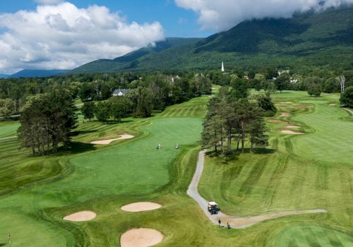 A scenic golf course with well-maintained greens, sand bunkers, and a golf cart on a path. Mountains in the background under a partly cloudy sky.