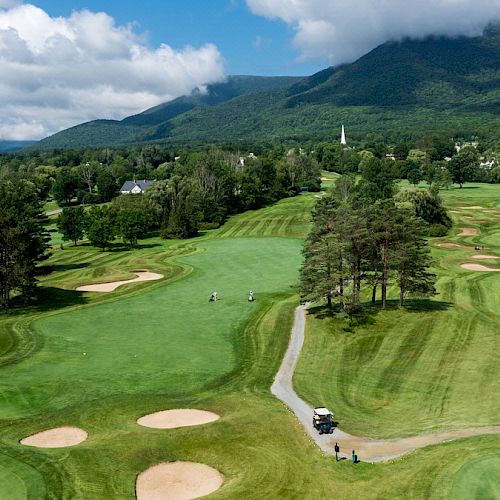 A scenic golf course with well-maintained greens, sand bunkers, and a golf cart on a path. Mountains in the background under a partly cloudy sky.