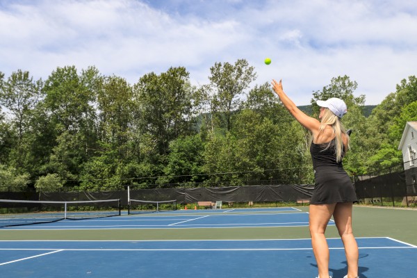 A woman in athletic wear prepares to serve a tennis ball on an outdoor court surrounded by trees and a blue sky.