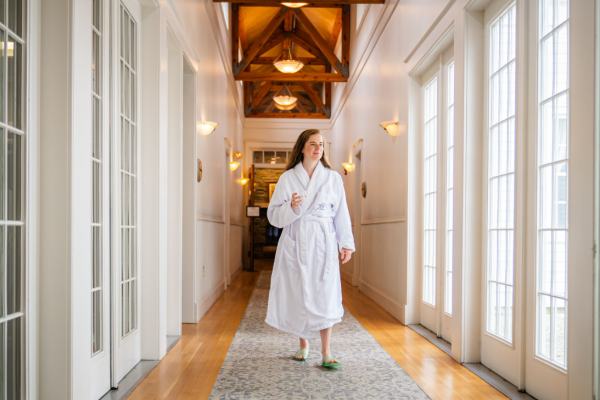 A person in a white robe walks down a well-lit corridor with wooden floors and ceiling beams, lined with windows on one side.