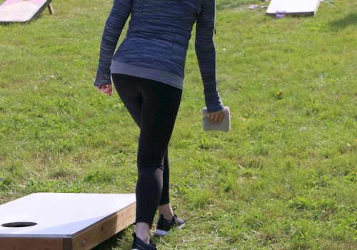 Two people are playing cornhole on a grassy area. One person is throwing a bean bag while the other waits at the opposite board.