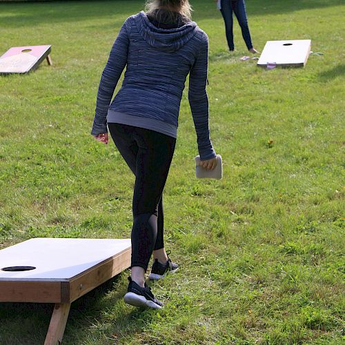 Two people are playing cornhole on a grassy area. One person is throwing a bean bag while the other waits at the opposite board.