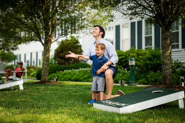 A man and child play cornhole on a grassy lawn, smiling and enjoying themselves.