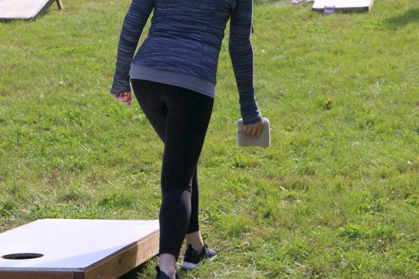 Two people playing cornhole on a grassy area, one wearing a gray hoodie and holding a bean bag, the other wearing a yellow shirt in the distance.