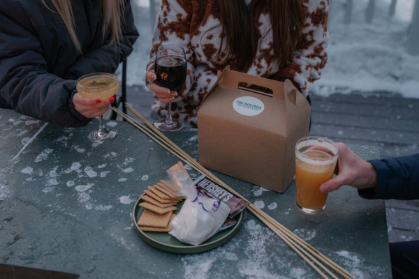 Three people are holding drinks around a table with graham crackers, marshmallows, chocolate, skewers, and a cardboard box labeled 