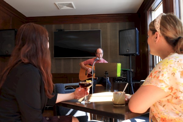 A man plays guitar and sings at a small venue, while two women enjoy drinks at a nearby table.