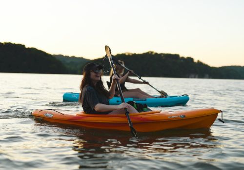 Two individuals are kayaking on a calm lake during sunset, with forested hills in the background, enjoying a peaceful outdoor adventure.