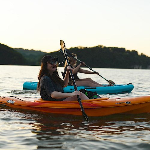 Two individuals are kayaking on a calm lake during sunset, with forested hills in the background, enjoying a peaceful outdoor adventure.