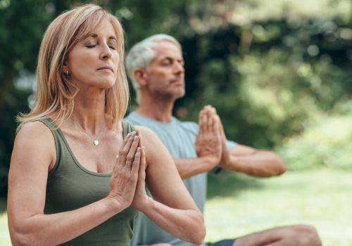 A woman and a man are meditating outdoors, sitting cross-legged with their hands in a prayer position and eyes closed.