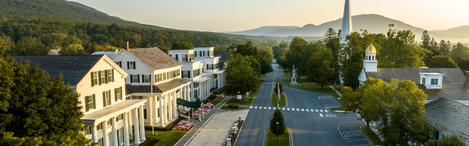 An aerial view of a small, picturesque town with white buildings, a church spire, and mountains in the background under clear skies.