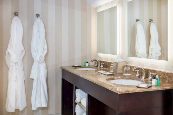 A bathroom with two hanging white robes, a dark wood vanity with two sinks, a marble countertop, mirrors, and toiletries on the counter.
