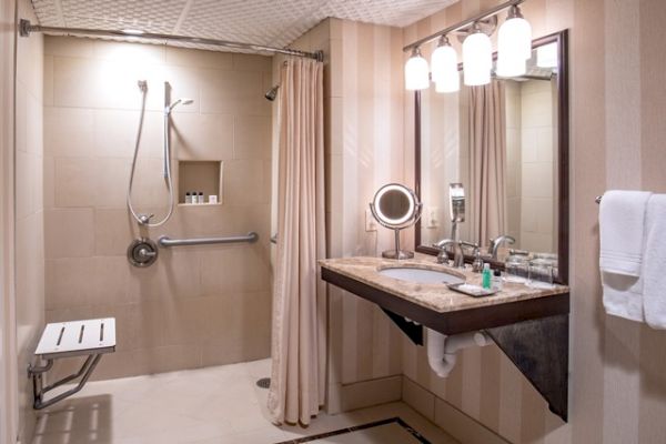 This image shows an accessible bathroom with a walk-in shower, grab bars, a foldable bench, a vanity with a mirror, and well-lit fixtures ending the sentence.