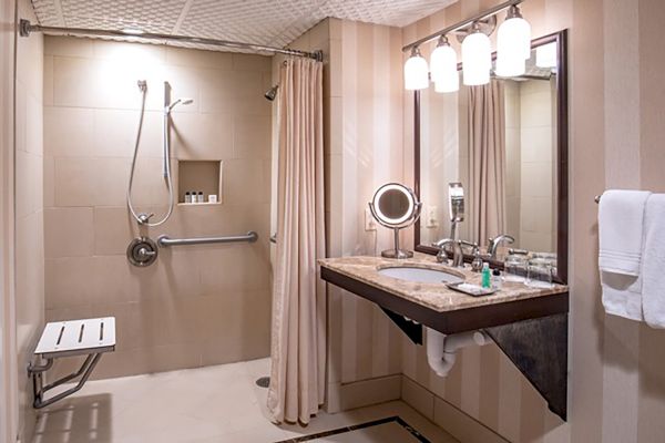 The image shows an accessible bathroom with a walk-in shower, bench, grab bar, and a vanity area with a sink, mirror, and toiletries under bright lights.