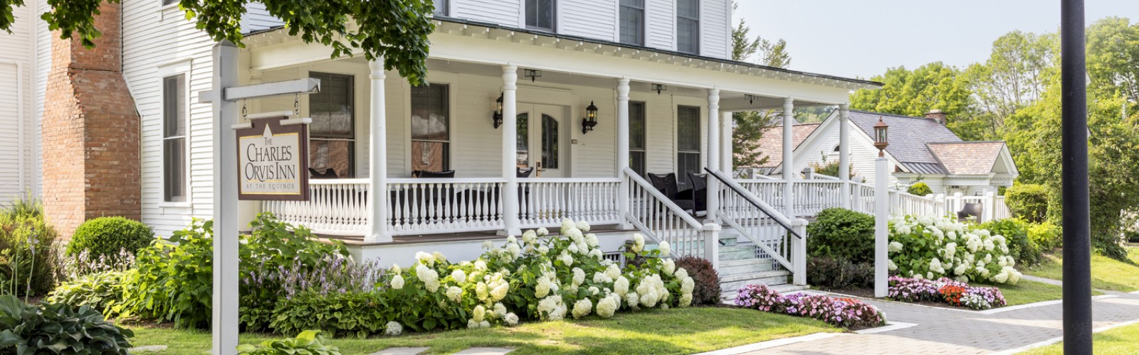 A charming, white two-story house with a porch, surrounded by lush greenery, flowers, and a streetlamp on a sunny day.