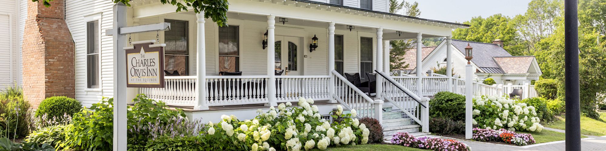 A charming, white two-story house with a porch, surrounded by lush greenery, flowers, and a streetlamp on a sunny day.