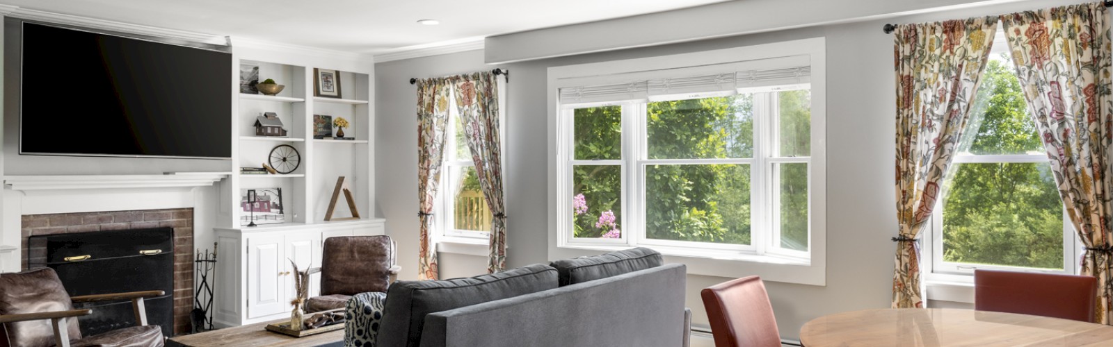 A bright living room with large windows, a gray sofa, a fireplace under a TV, bookshelves, a colorful rug, and a round dining table with chairs.