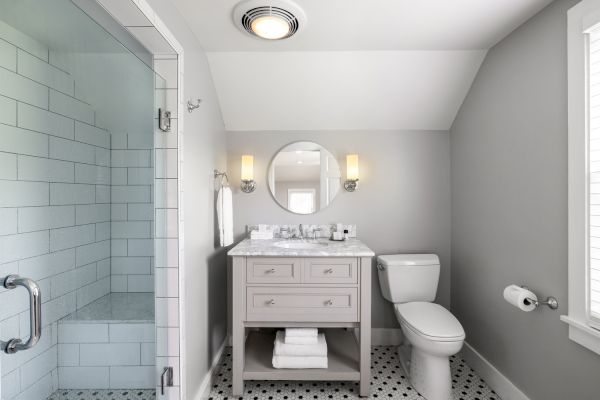 A modern bathroom features a glass-enclosed shower, a marble-top vanity with a round mirror, wall sconces, and a toilet next to a window.