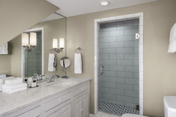 A modern bathroom with a gray tiled shower, a marble countertop with two sinks, a mirror, and folded towels on a counter.