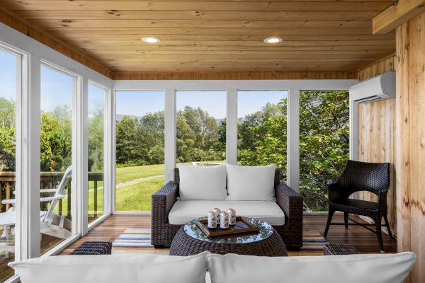 A cozy sunroom features wicker furniture with white cushions, a round glass coffee table, and panoramic windows offering a view of a lush green landscape.