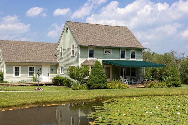 A light green house with a front porch, lawn, and adjacent pond, set against a blue sky with clouds.