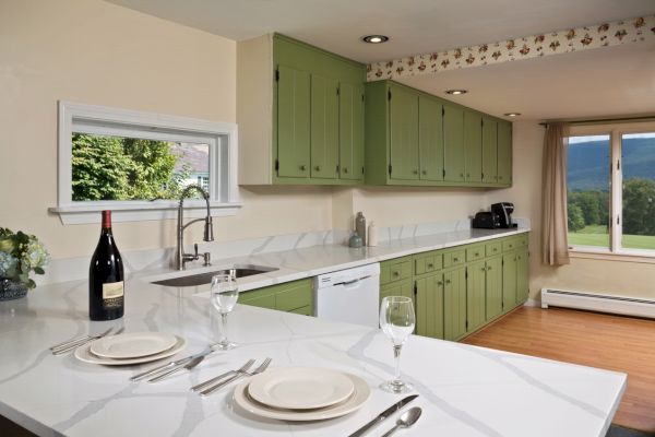 A well-lit kitchen with green cabinets, white countertops, and a window view. It has a dining setup with wine, plates, and cutlery on the counter.