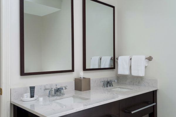 A clean bathroom features a double sink, two mirrors, and towel racks with white towels, along with a tissue box and toiletries on the counter.