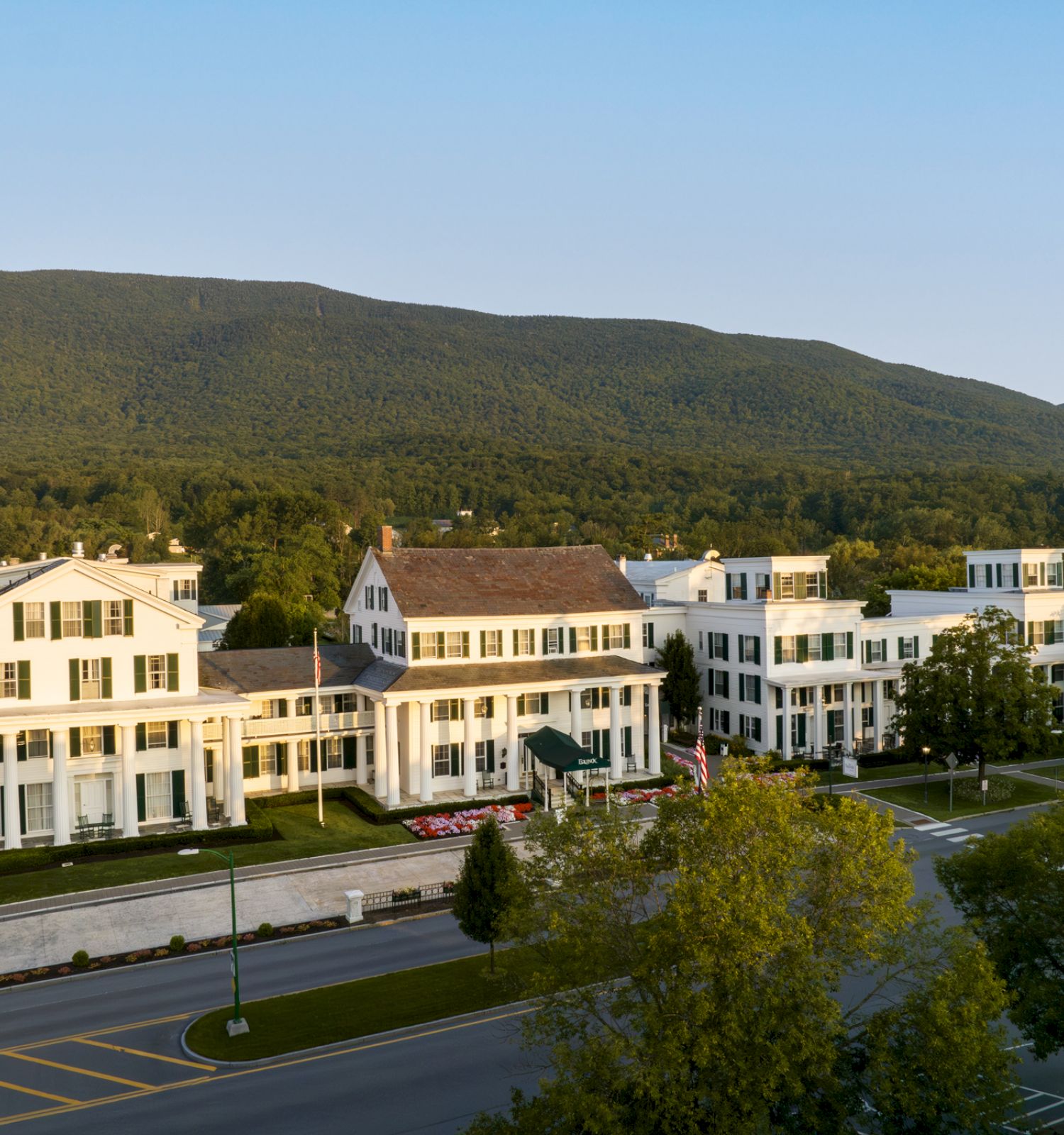 An expansive building with white facades set against a backdrop of a lush, green mountain and trees, intersected by a road in the foreground.