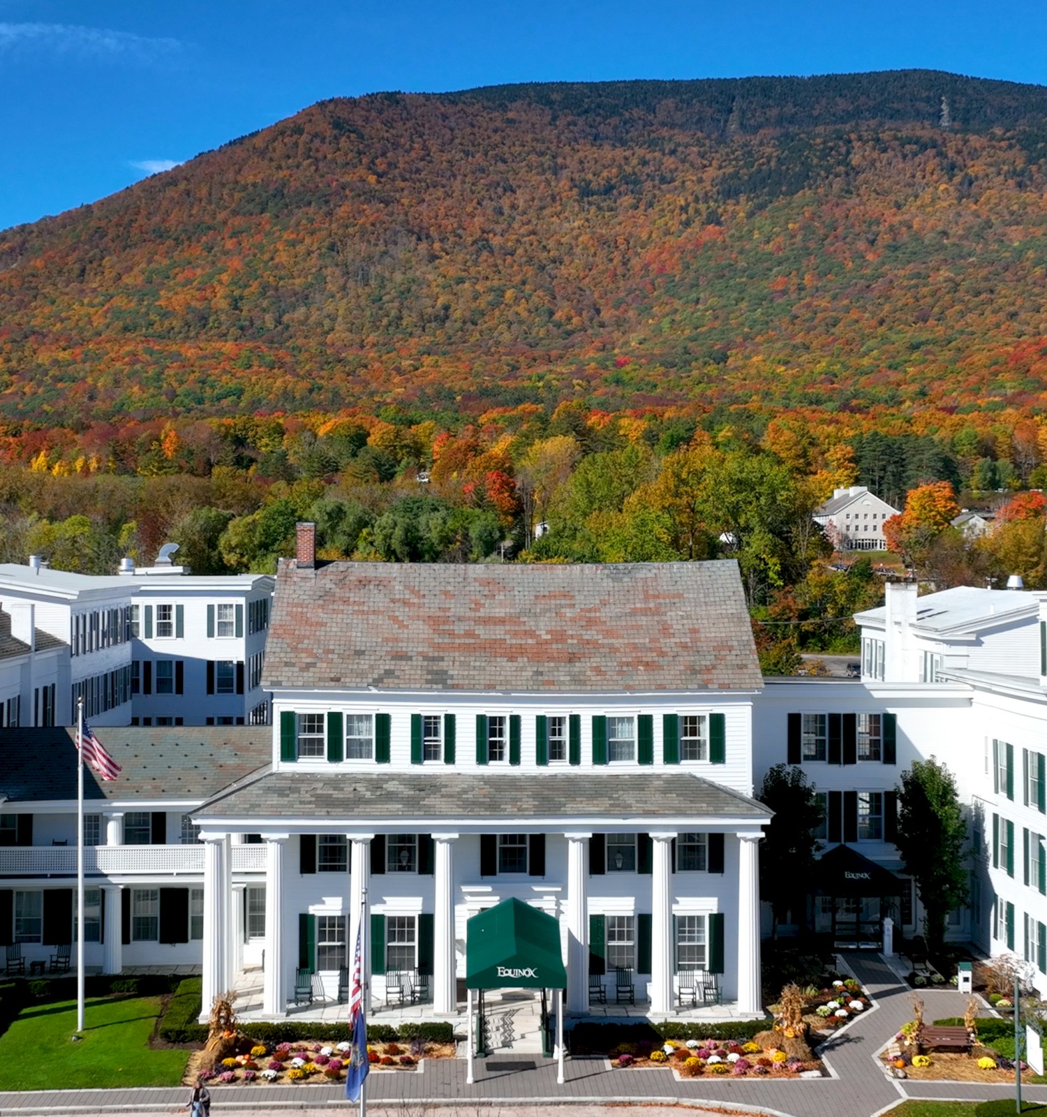 The image shows a large white building with green shutters, surrounded by autumn foliage and mountains in the background.