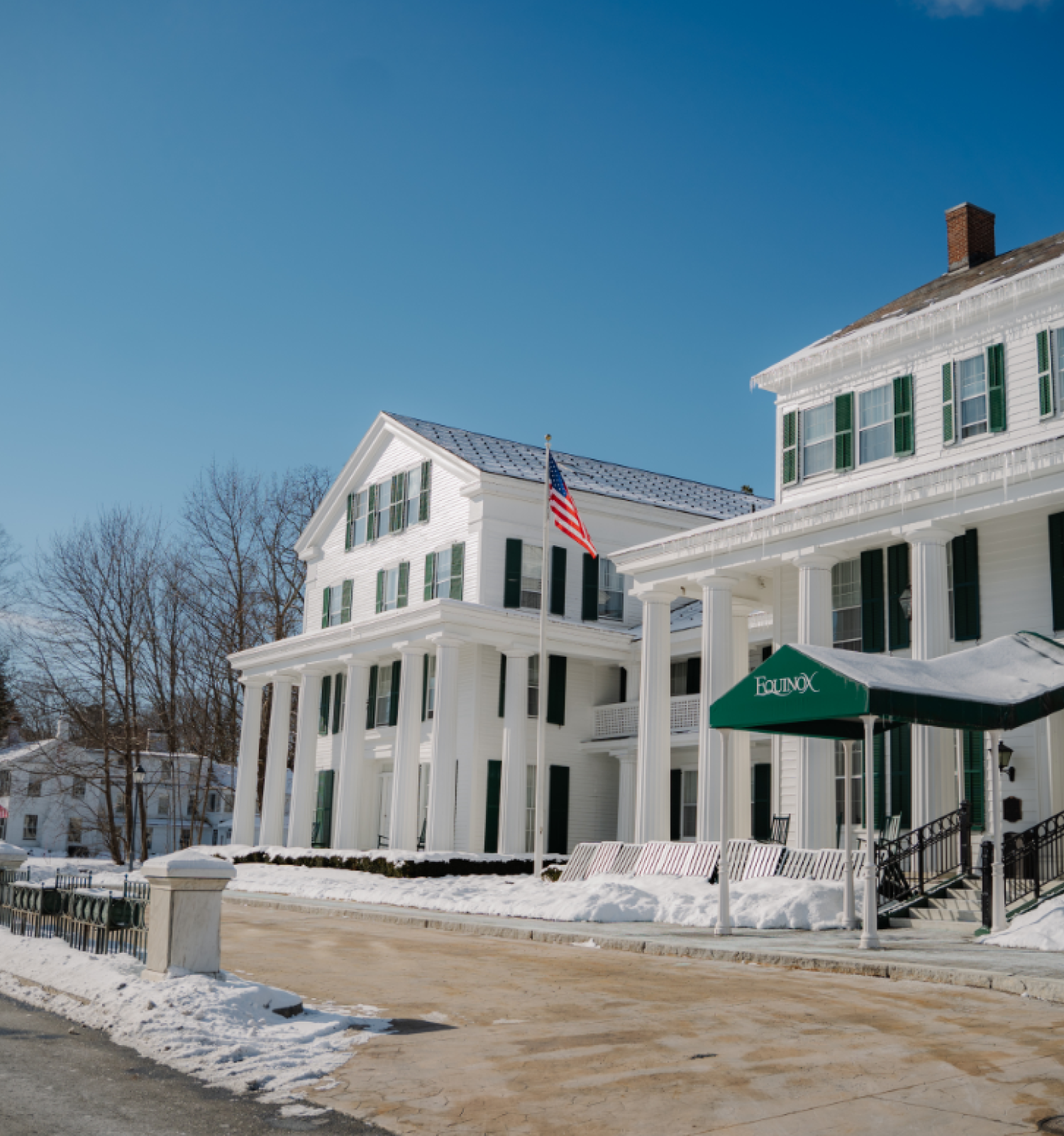 A large white building with green shutters and an American flag, surrounded by snow, under a clear blue sky.